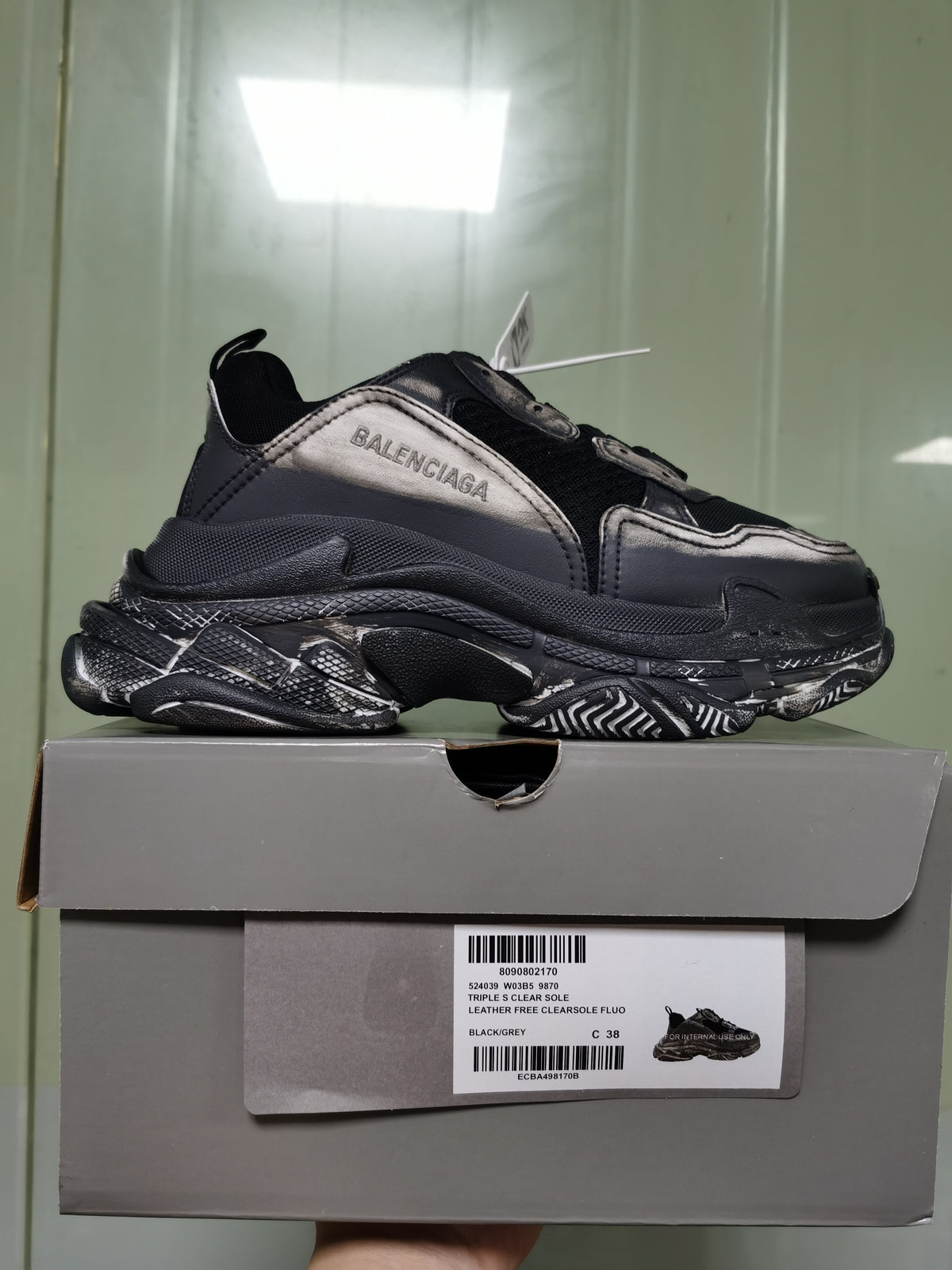 Side profile of the sneaker displayed on its box; the box label identifies Triple S Clear Sole, style code 9870, black/grey, and size C 38.