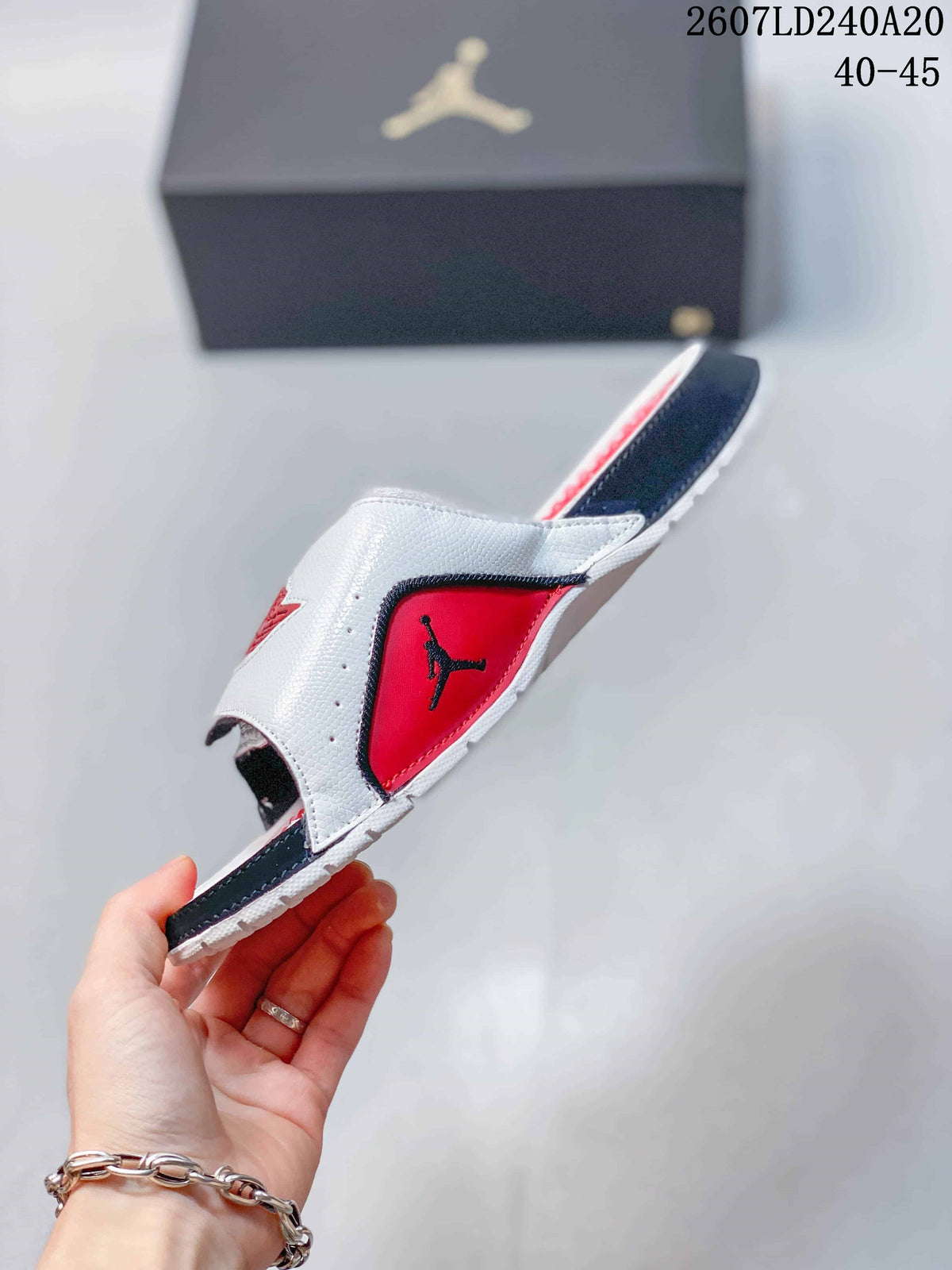 Side view of a white Jordan slide with red and black panels, adjustable strap, and patterned outsole; image text shows style code 2607LD240A20 and sizes 40–45.