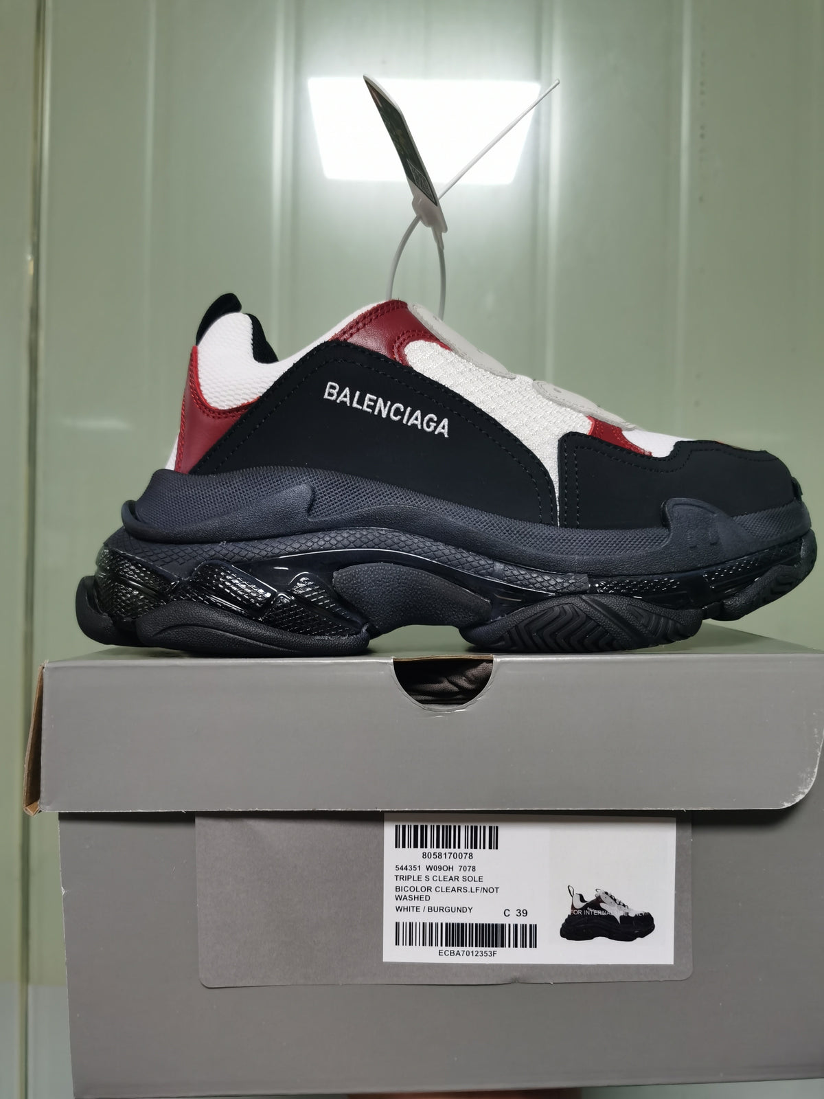 Side view of a Balenciaga Triple S sneaker in white, burgundy, black and dark grey, displayed on its labeled box.