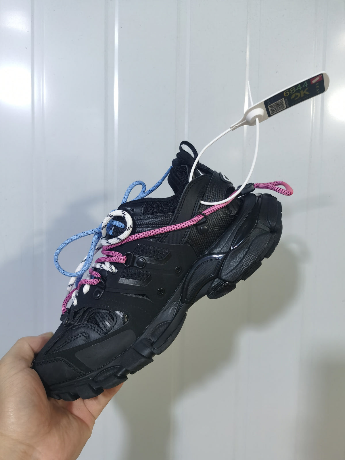 Black Balenciaga Track Trail sneaker shown from an angled side view with layered panels, chunky sole, and pink and blue rope-style laces.