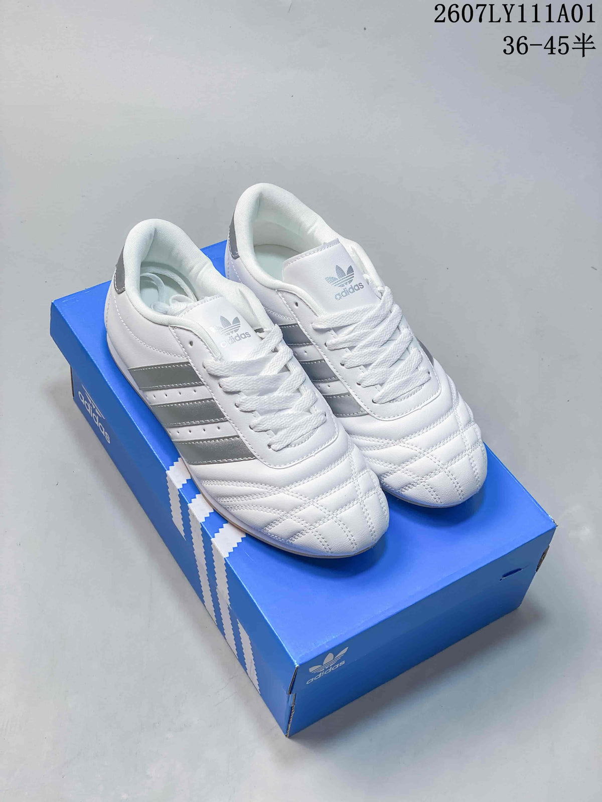 Pair of white Adidas Taekwondo low-top sneakers with silver stripes and quilted toes on a blue shoe box.