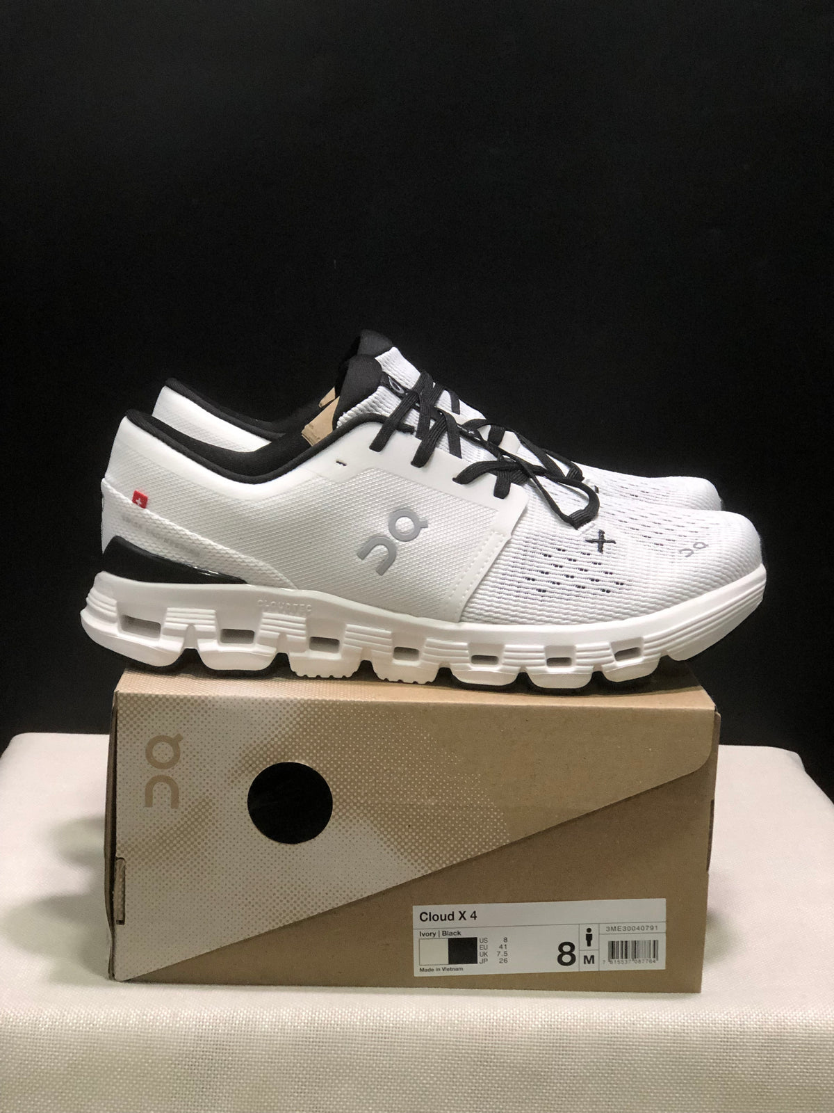 Side view of an Ivory and Black On Cloud X4 sneaker with the shoe box label visible.