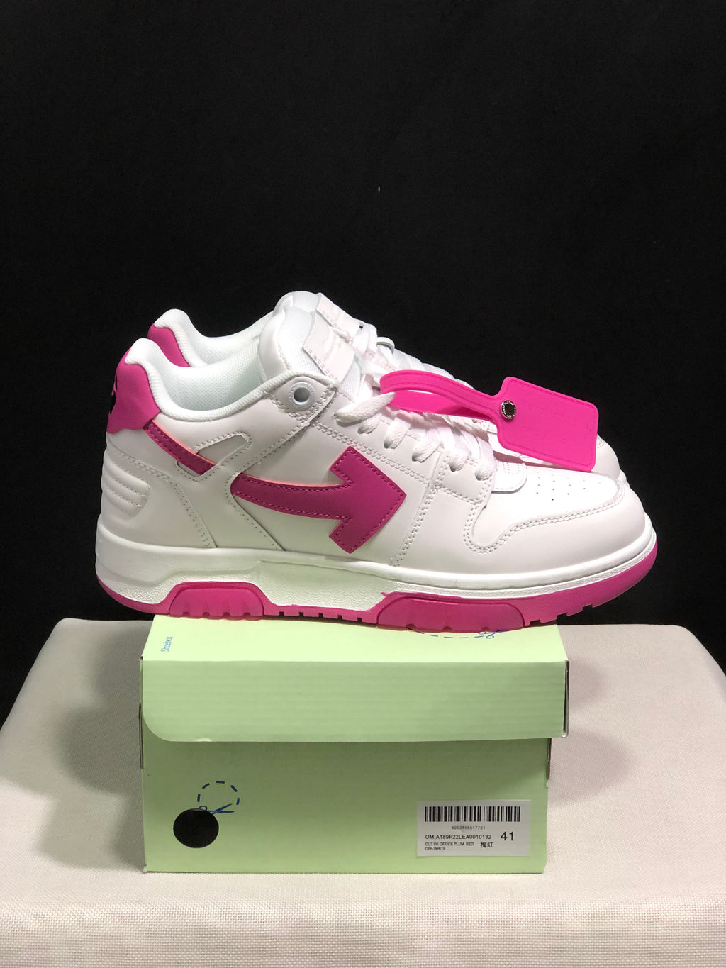 Side view of a white sneaker with a pink arrow-shaped side detail, pink outsole accents and a detachable tag.
