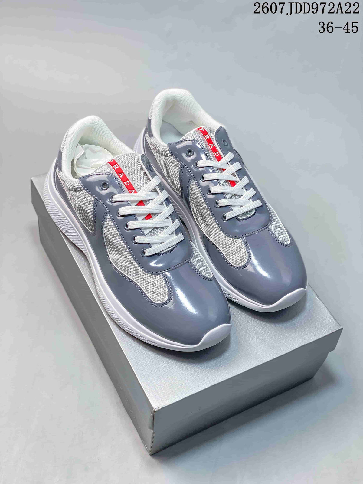 Pair of gray and white Prada America's Cup low-top sneakers displayed on a box, with red tongue branding.