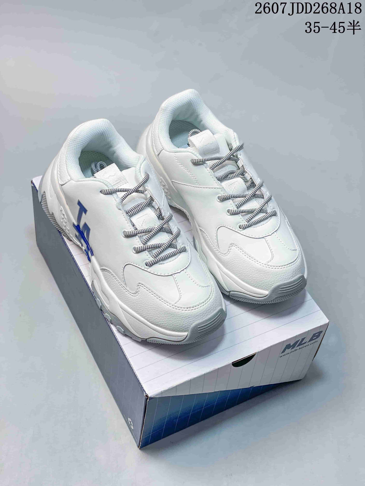 Pair of white MLB low-top sneakers with blue NY side lettering, patterned laces and a chunky outsole displayed on an MLB shoe box.