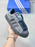 Close-up angled view of a gray and black adidas sneaker highlighting the trefoil tongue logo, three-stripe panel, laces, shell toe, and dark sole.