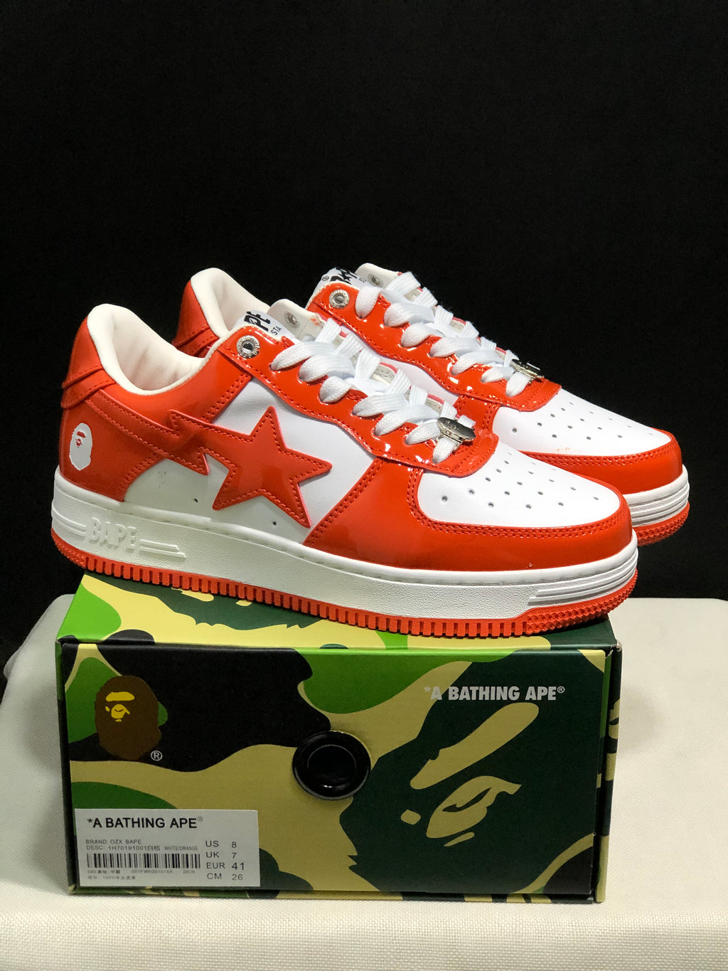 White and orange BAPE Sta sneakers displayed on a camouflage BAPE shoebox; the box label shows a photographed sample in EU 41 and CM 26.
