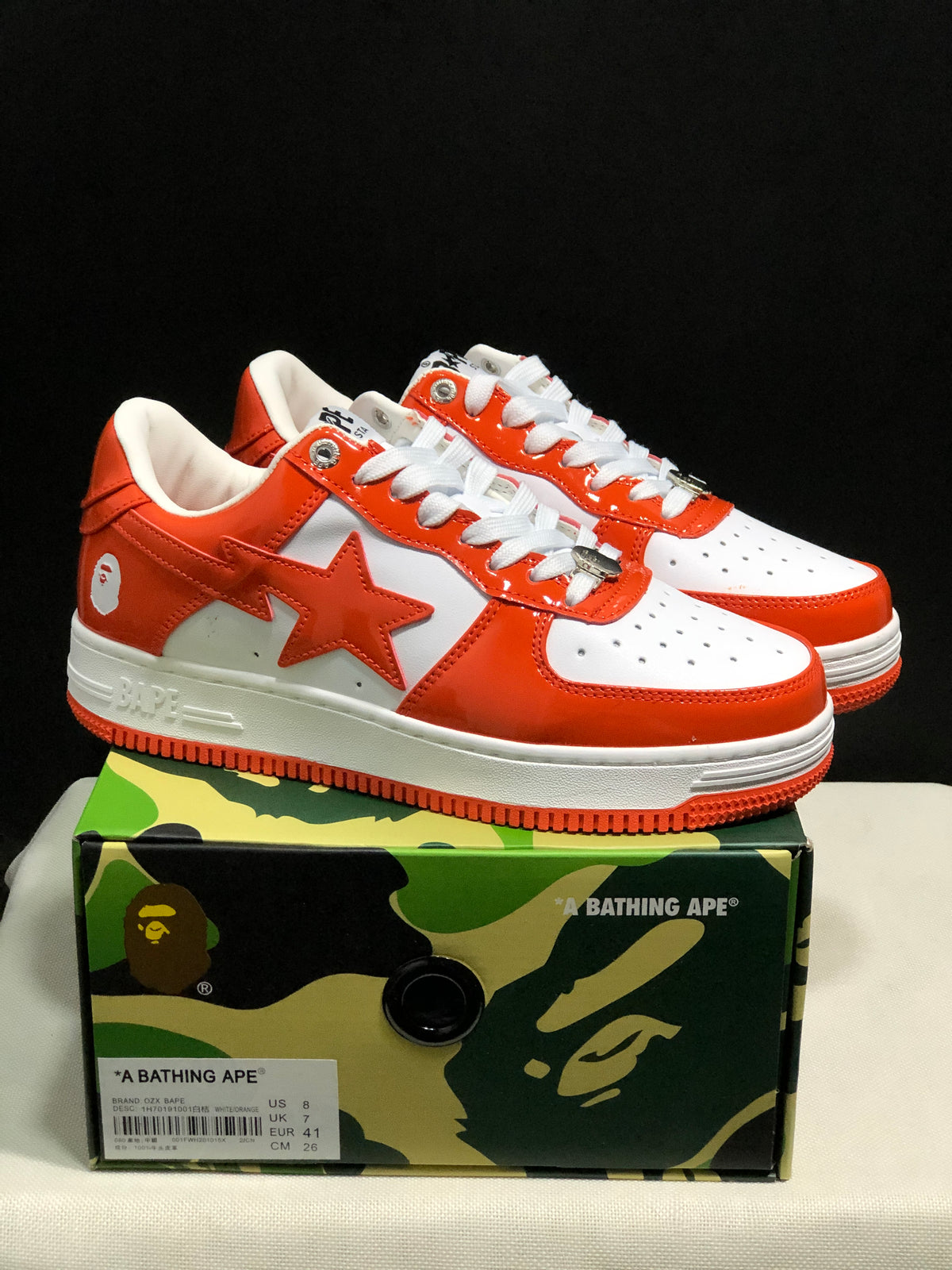 White and orange BAPE Sta sneakers displayed on a camouflage BAPE shoebox; the box label shows a photographed sample in EU 41 and CM 26.