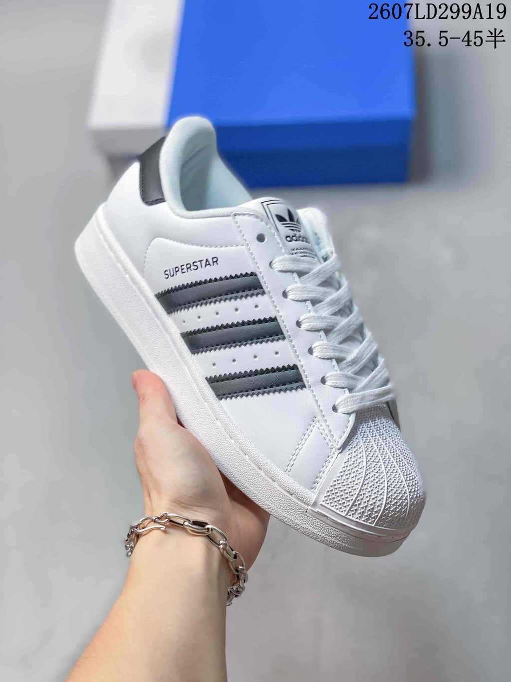 Angled side view of a white adidas Superstar sneaker with black stripes, shell toe, white sole and visible SUPERSTAR marking.