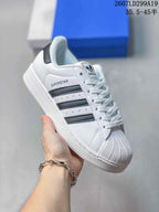 Angled side view of a white adidas Superstar sneaker with black stripes, shell toe, white sole and visible SUPERSTAR marking.