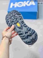 Close-up of the dark lugged outsole with visible Vibram branding; a HOKA shoebox and size overlay appear in the background.