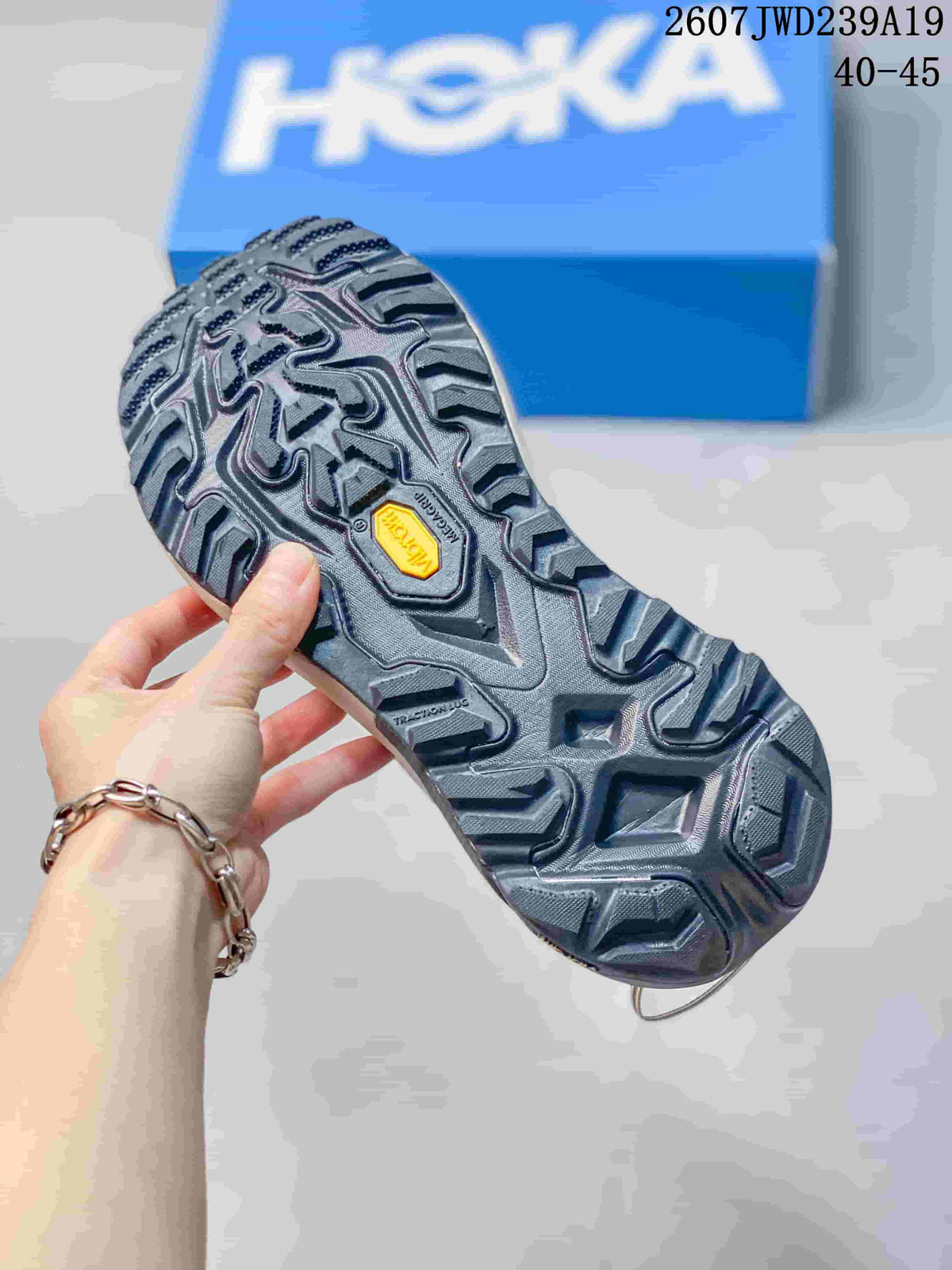 Close-up of the dark lugged outsole with visible Vibram branding; a HOKA shoebox and size overlay appear in the background.