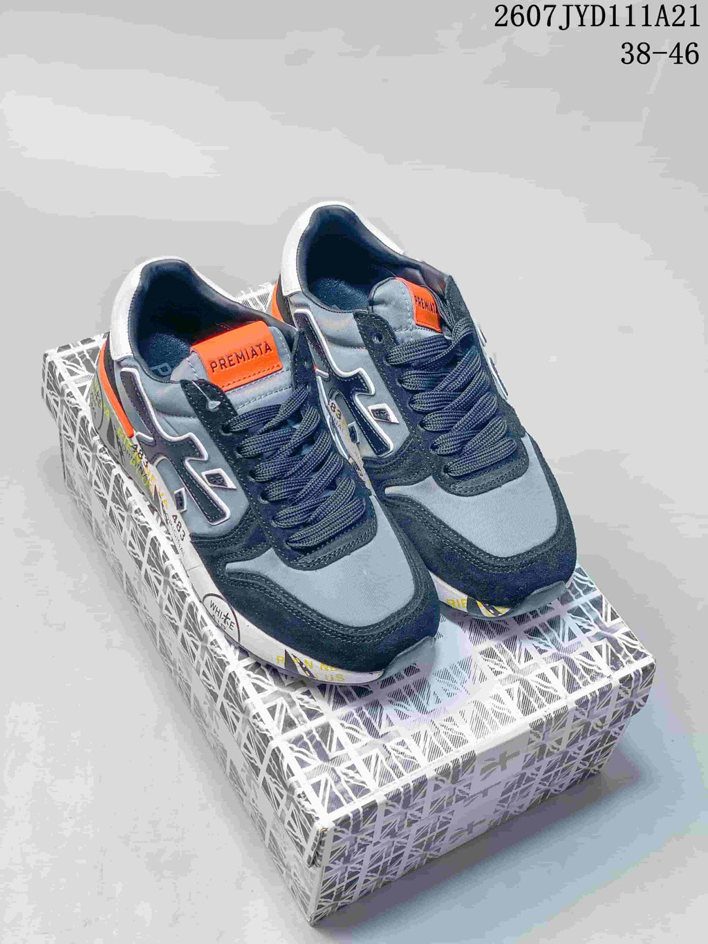 Pair of grey and navy Premiata sneakers with orange accents displayed on a patterned shoebox.