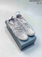 Pair of pale gray Onitsuka Tiger sneakers with broad ribbon-style laces and textured textile uppers on a shoebox.