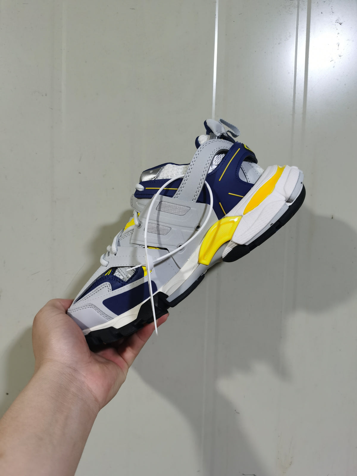 Balenciaga Track sneaker held at an angle, showing white, navy blue and yellow layered panels with a black outsole.
