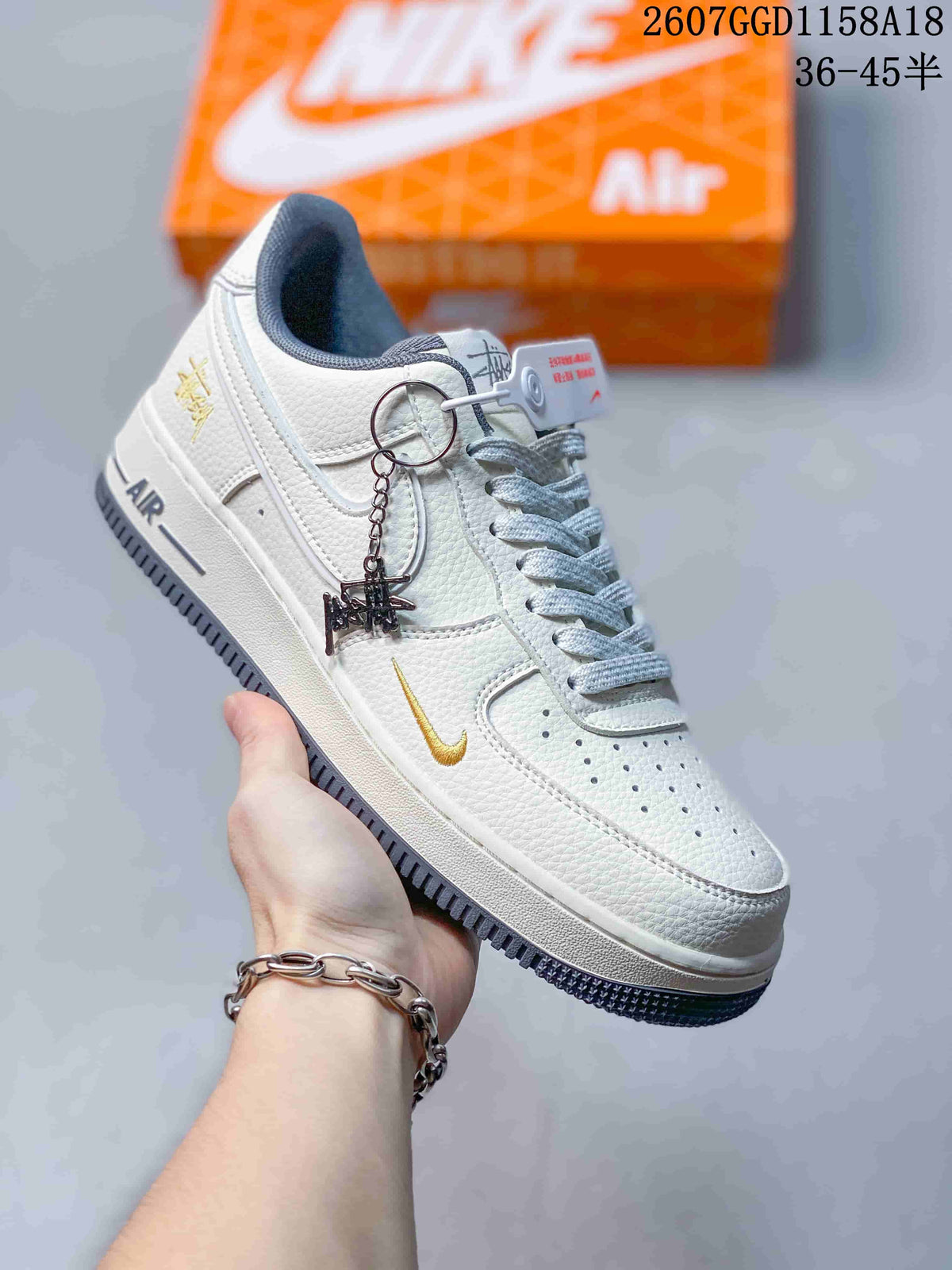Close-up side view of a white low-top sneaker with a contrasting swoosh, perforated toe, dark outsole, decorative side branding, and a lace accessory.