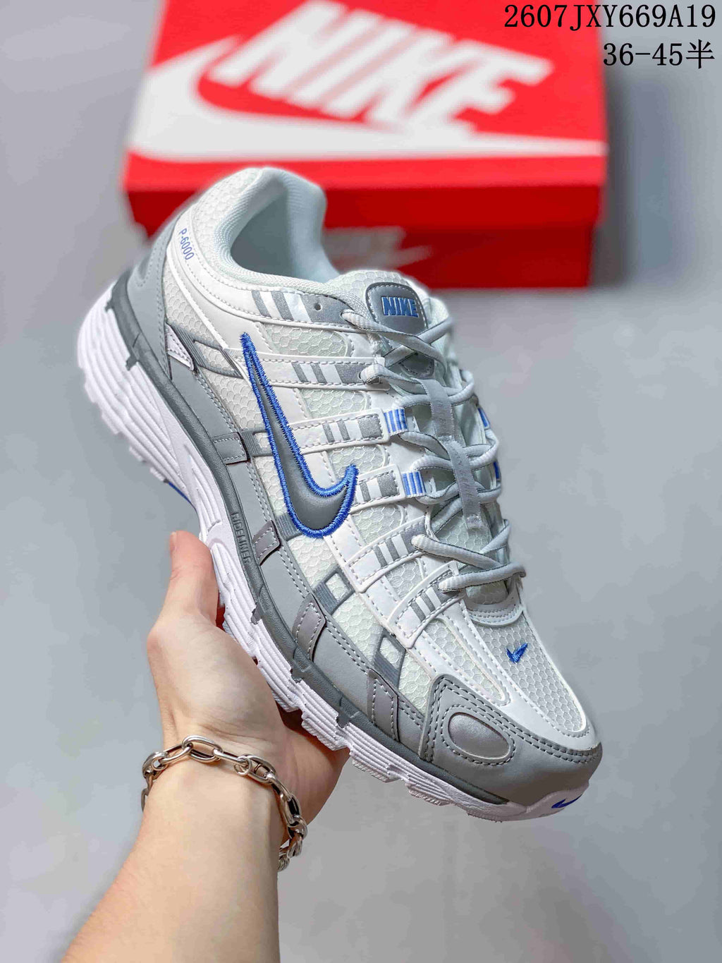 Angled side view of the white and grey sneaker with a blue Swoosh, padded collar, layered panels, and red shoebox in the background.