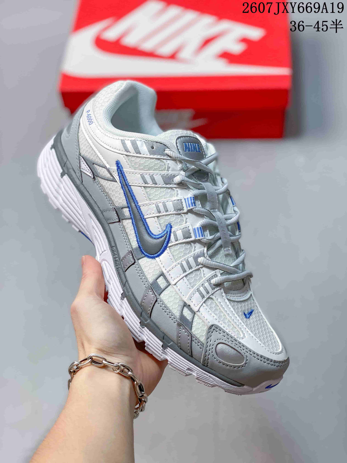 Angled side view of the white and grey sneaker with a blue Swoosh, padded collar, layered panels, and red shoebox in the background.