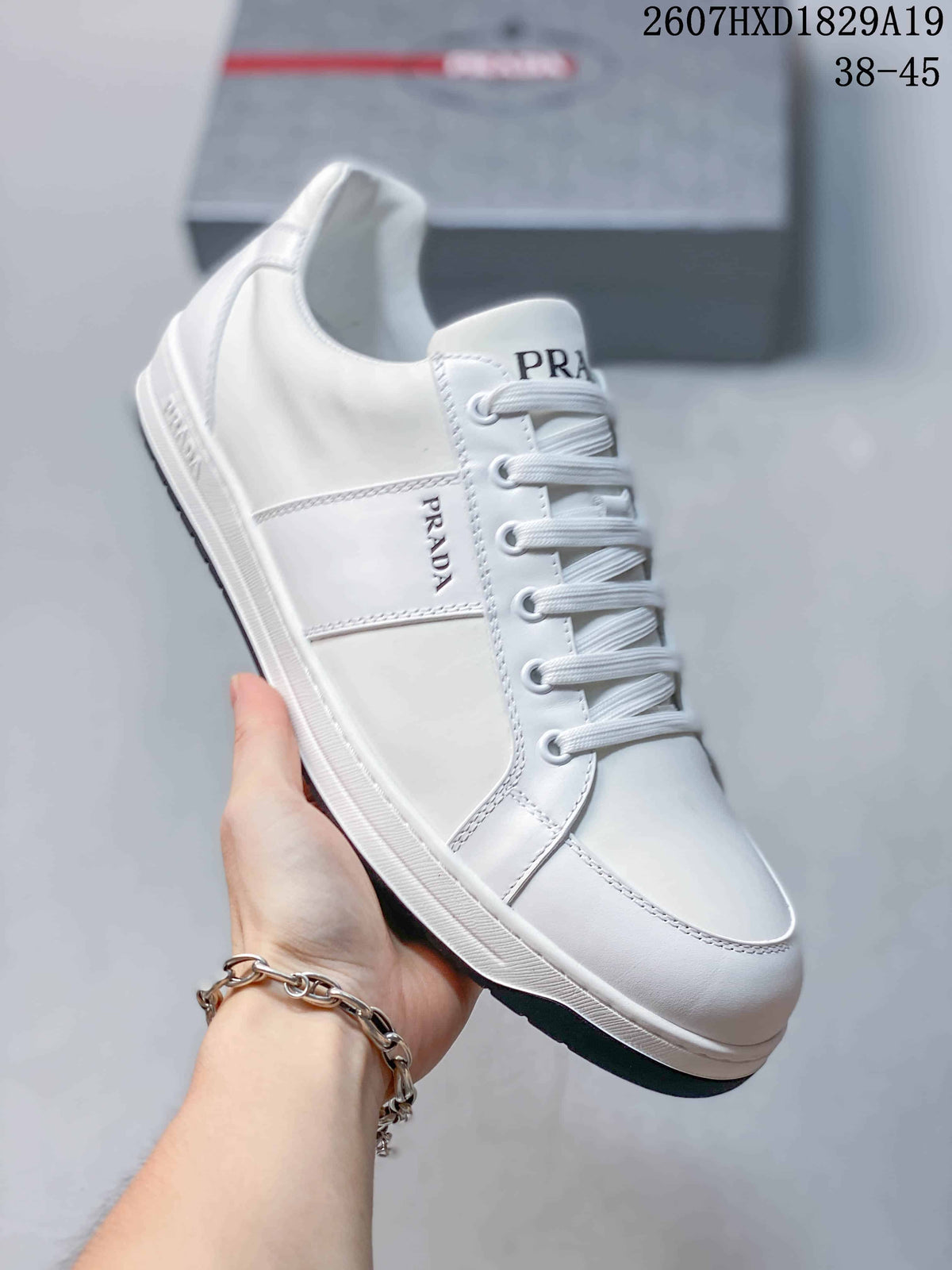 Side view of a white Prada Downtown low-top sneaker showing the lace-up front, branded side panel and light-colored sole; style code 2607HXD1829A19 and size range 38–45 are visible.