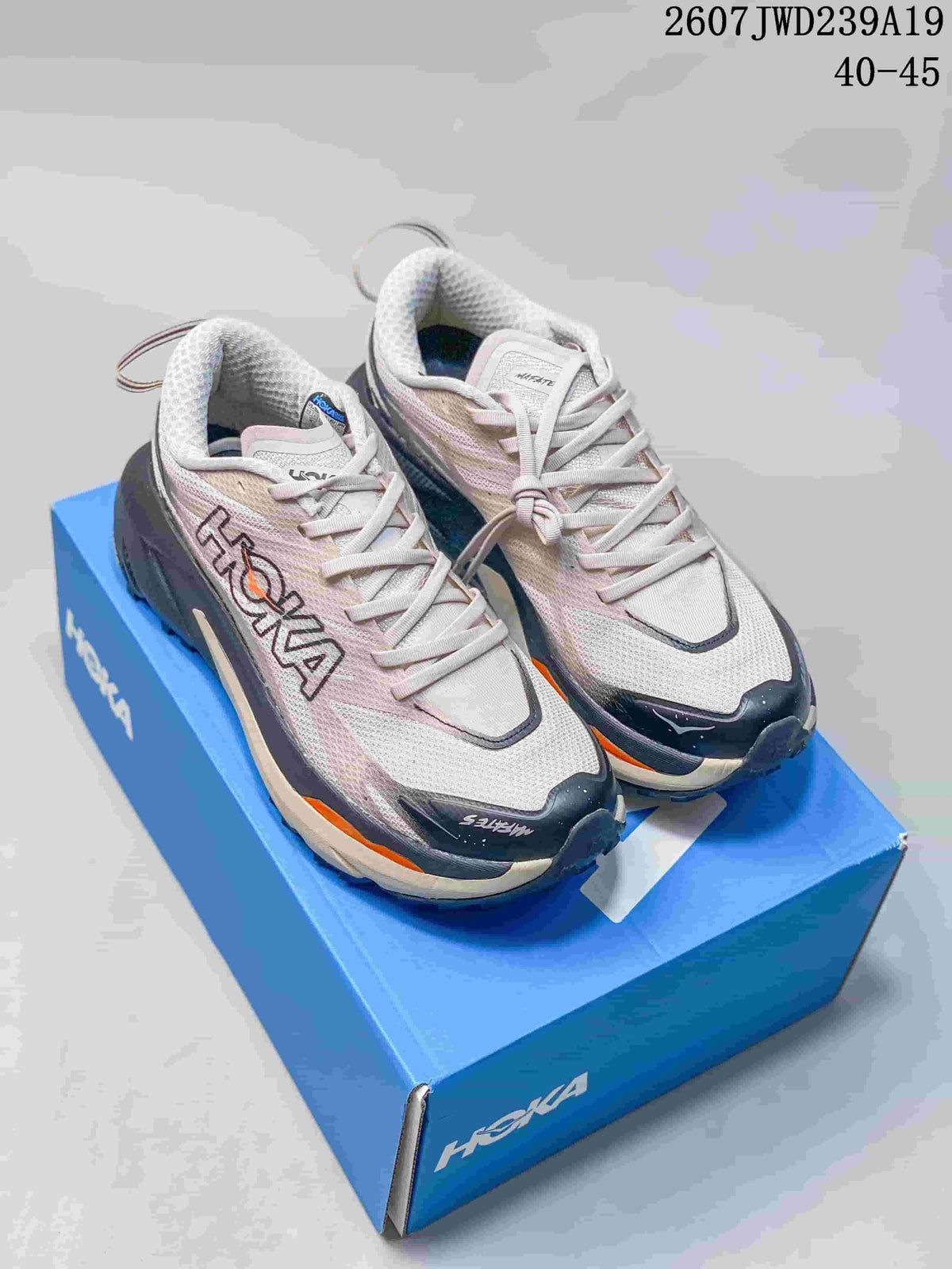 Pair of light-colored HOKA running shoes with navy and orange accents displayed on a blue shoebox; overlay text shows style code 2607JWD239A19 and size range 40–45.