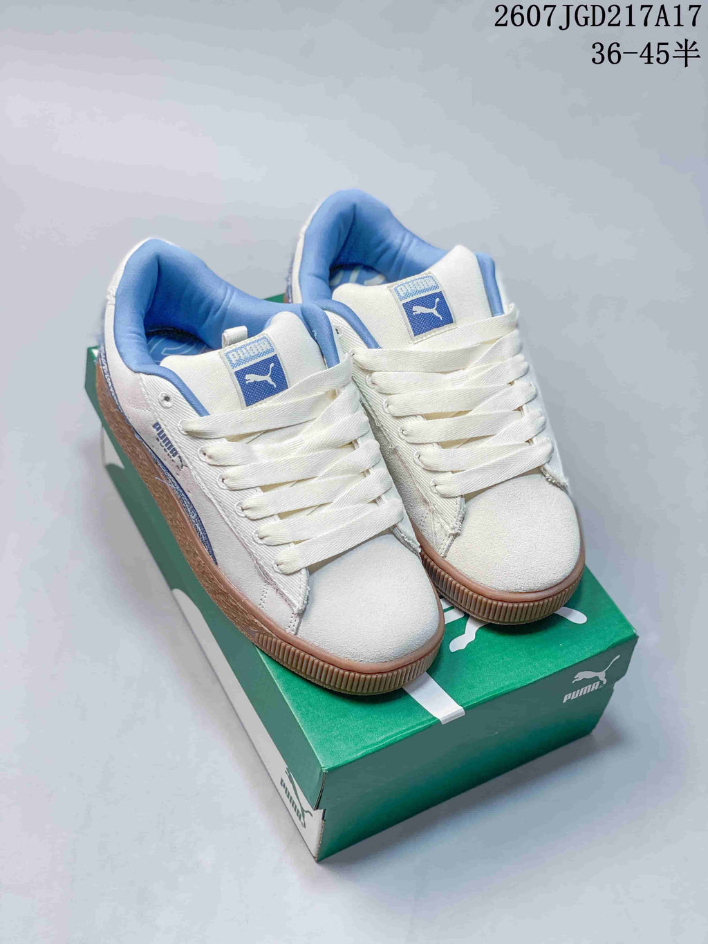 Pair of white PUMA low-top sneakers with blue accents and gum-colored soles displayed on a green PUMA shoe box; image text shows style code 2607JGD217A17 and size range 36–45.5.