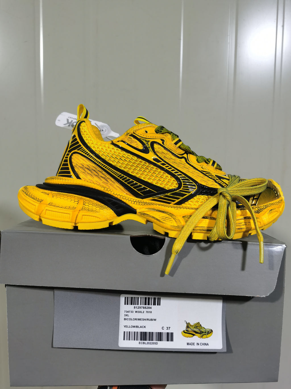 Side profile of the sneaker resting on a gray shoe box; the box label shows yellow/black color details and size C 37.