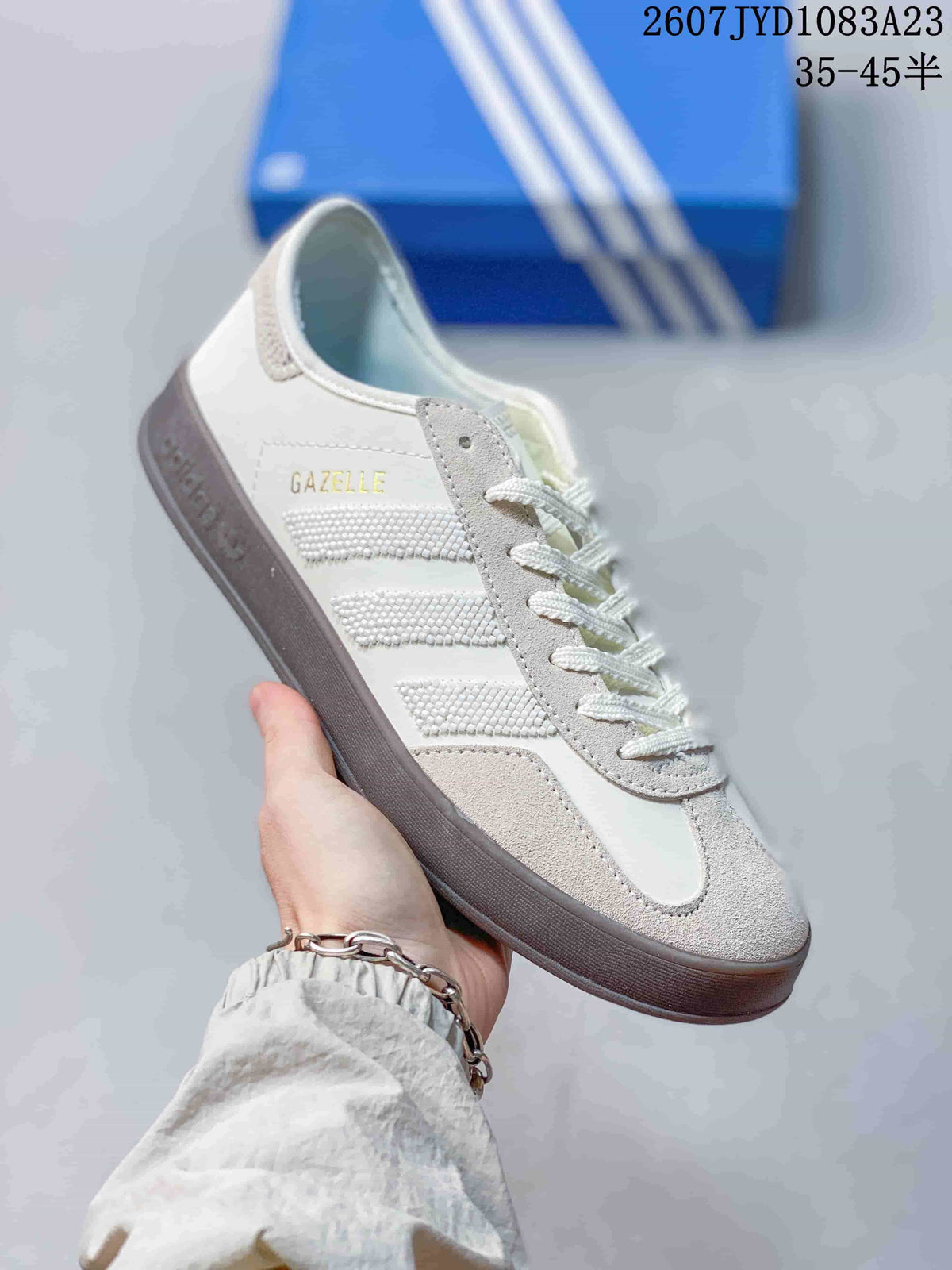 Angled side view of the white and gray sneaker showing the Gazelle marking, textured stripes, and dark outsole.