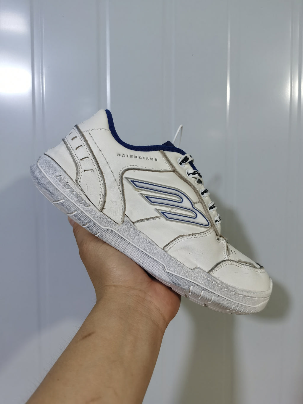 Side view of a white Balenciaga Hampton sneaker showing the blue-trimmed side emblem, stitched panels, and textured sole.