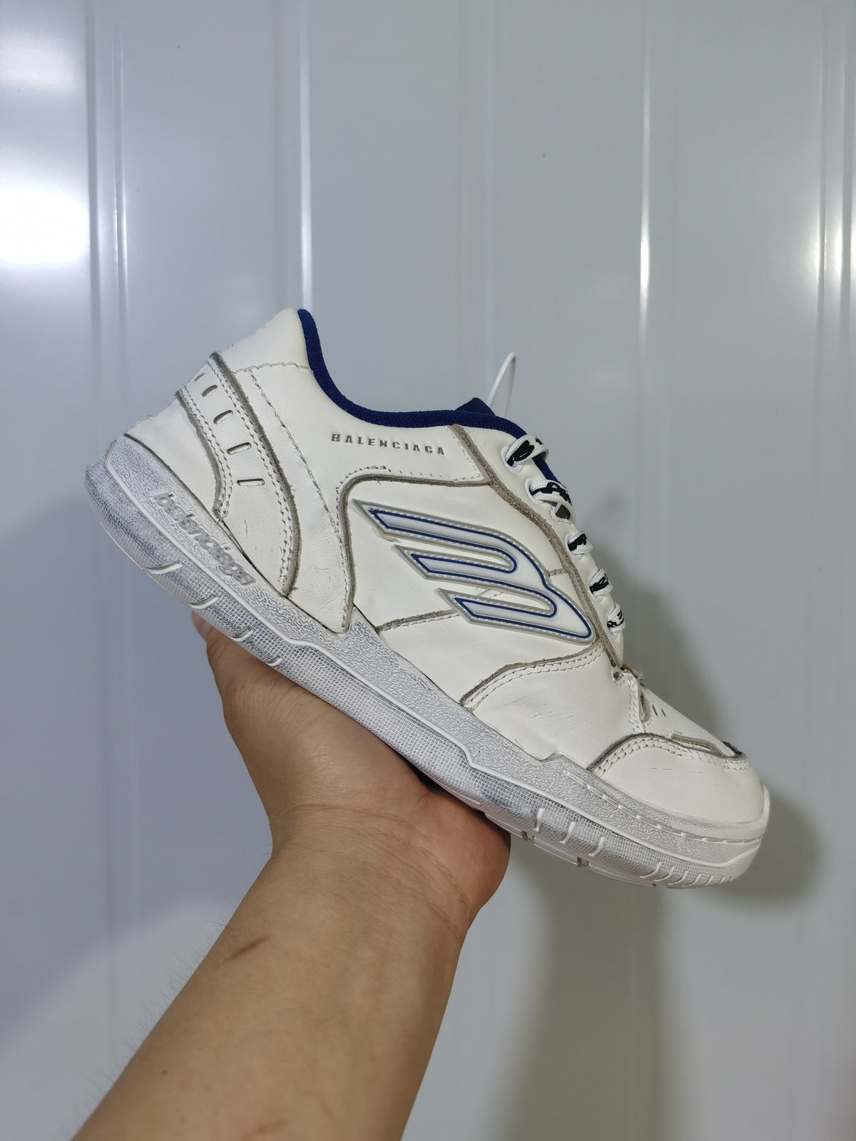 Side view of a white Balenciaga Hampton sneaker showing the blue-trimmed side emblem, stitched panels, and textured sole.
