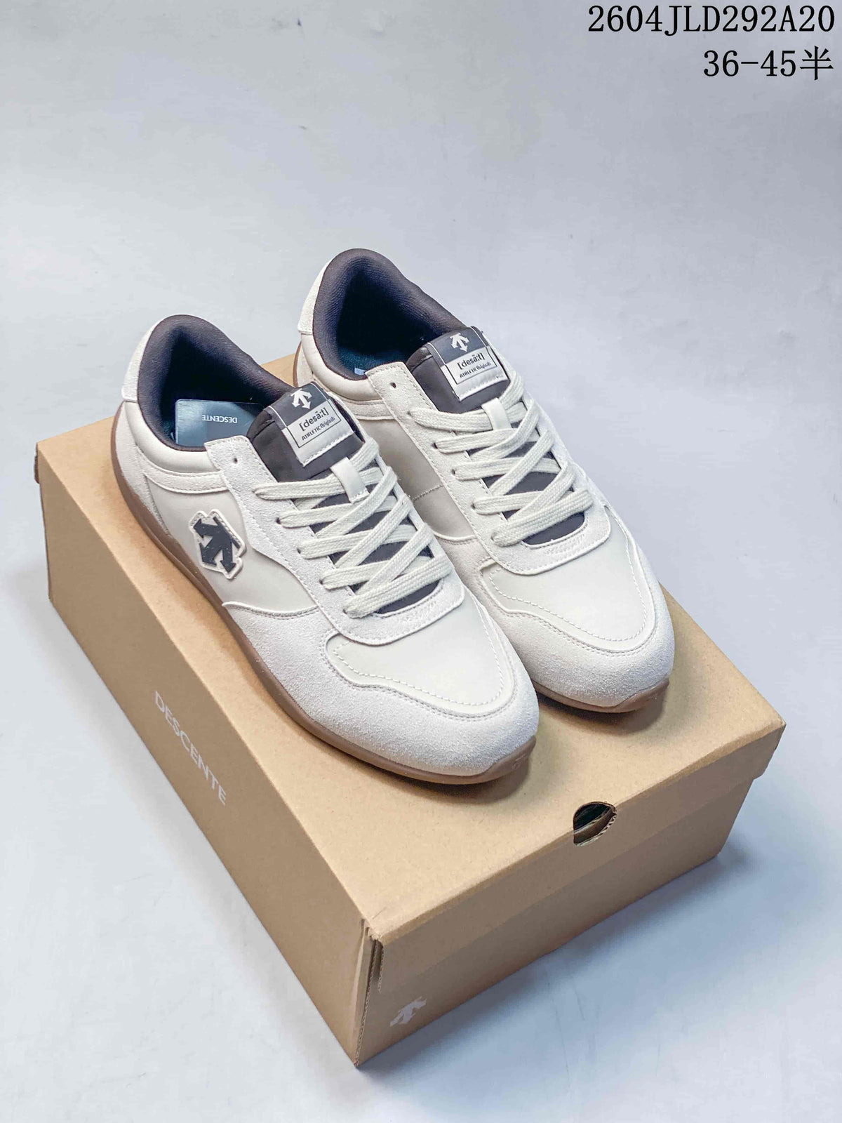 Pair of cream Descente Chron Tempo Slim low-top sneakers with gum-colored soles, shown on a shoe box.