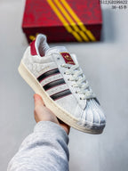Angled side view showing the Superstar profile, textured white upper, black stripes, red accents, and decorative charm.