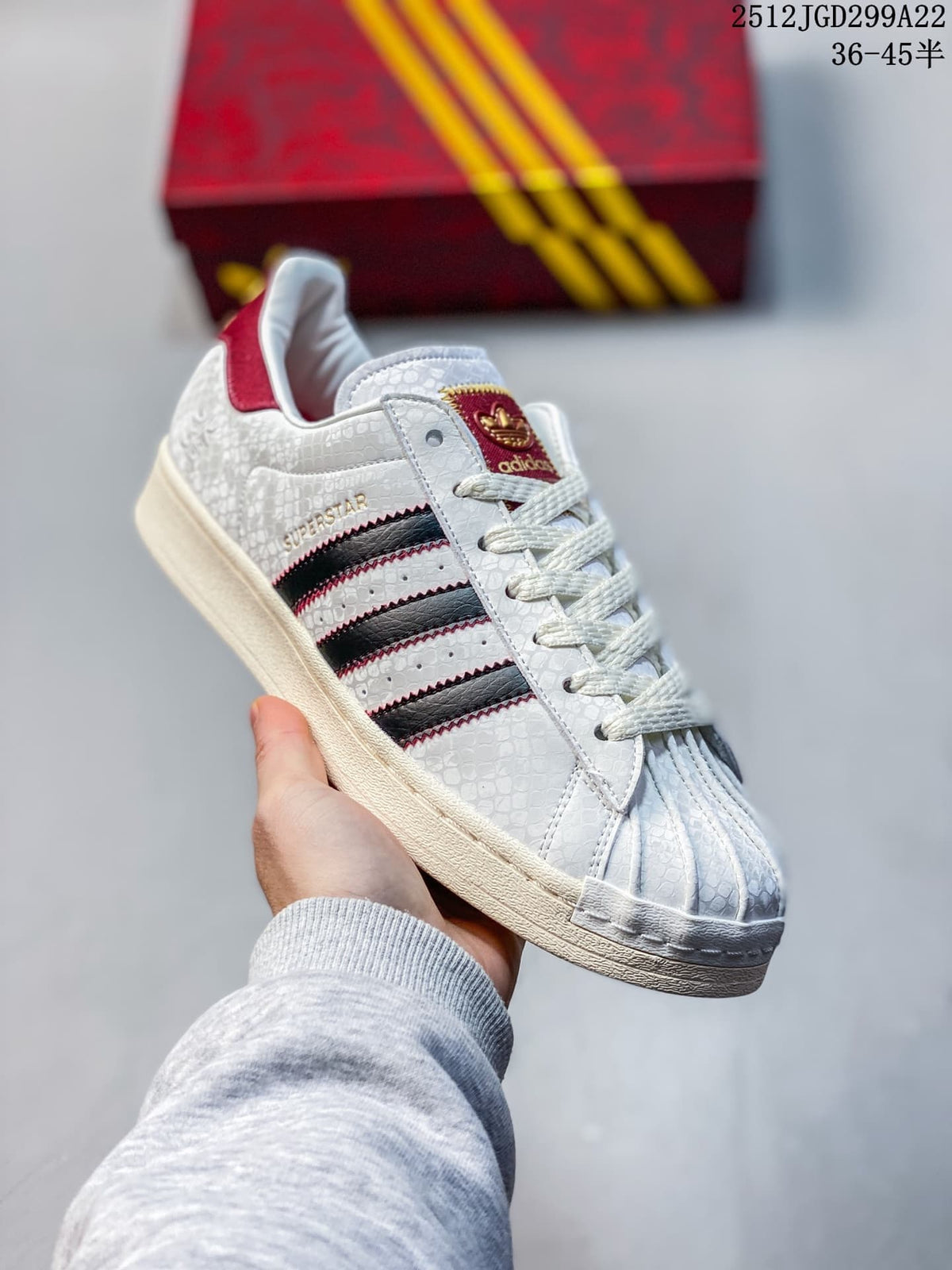 Angled side view showing the Superstar profile, textured white upper, black stripes, red accents, and decorative charm.