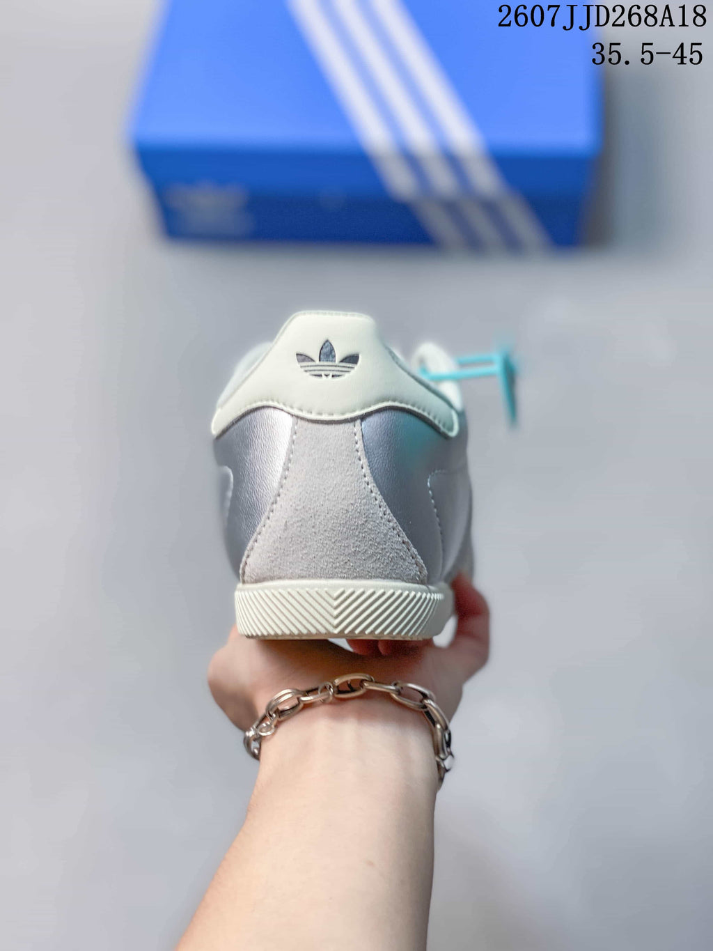 Rear view of a silver and white sneaker showing the heel panel, trefoil branding, and textured outsole edge.