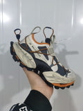 Close-up of a beige, black, and orange Balenciaga sneaker showing layered panels, cord-style lacing, gray webbing, and a sculpted sole.