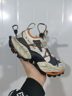 Close-up of a beige, black, and orange Balenciaga sneaker showing layered panels, cord-style lacing, gray webbing, and a sculpted sole.
