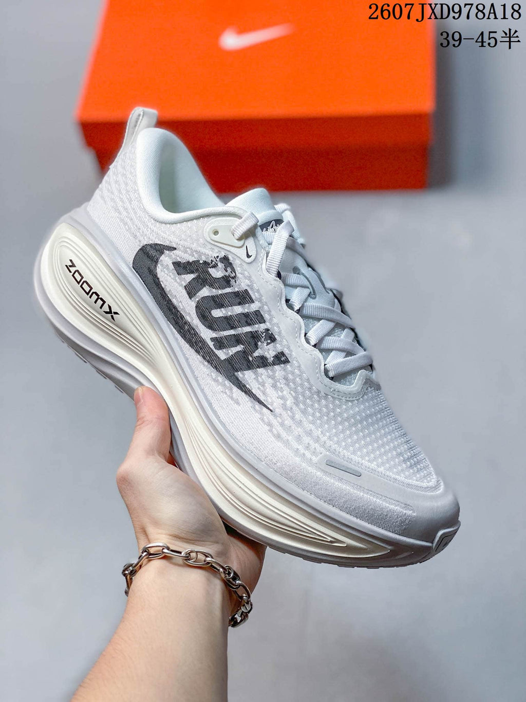 White Nike Vomero Plus running shoe with a light gray upper and visible ZoomX heel branding, photographed beside an orange shoe box.