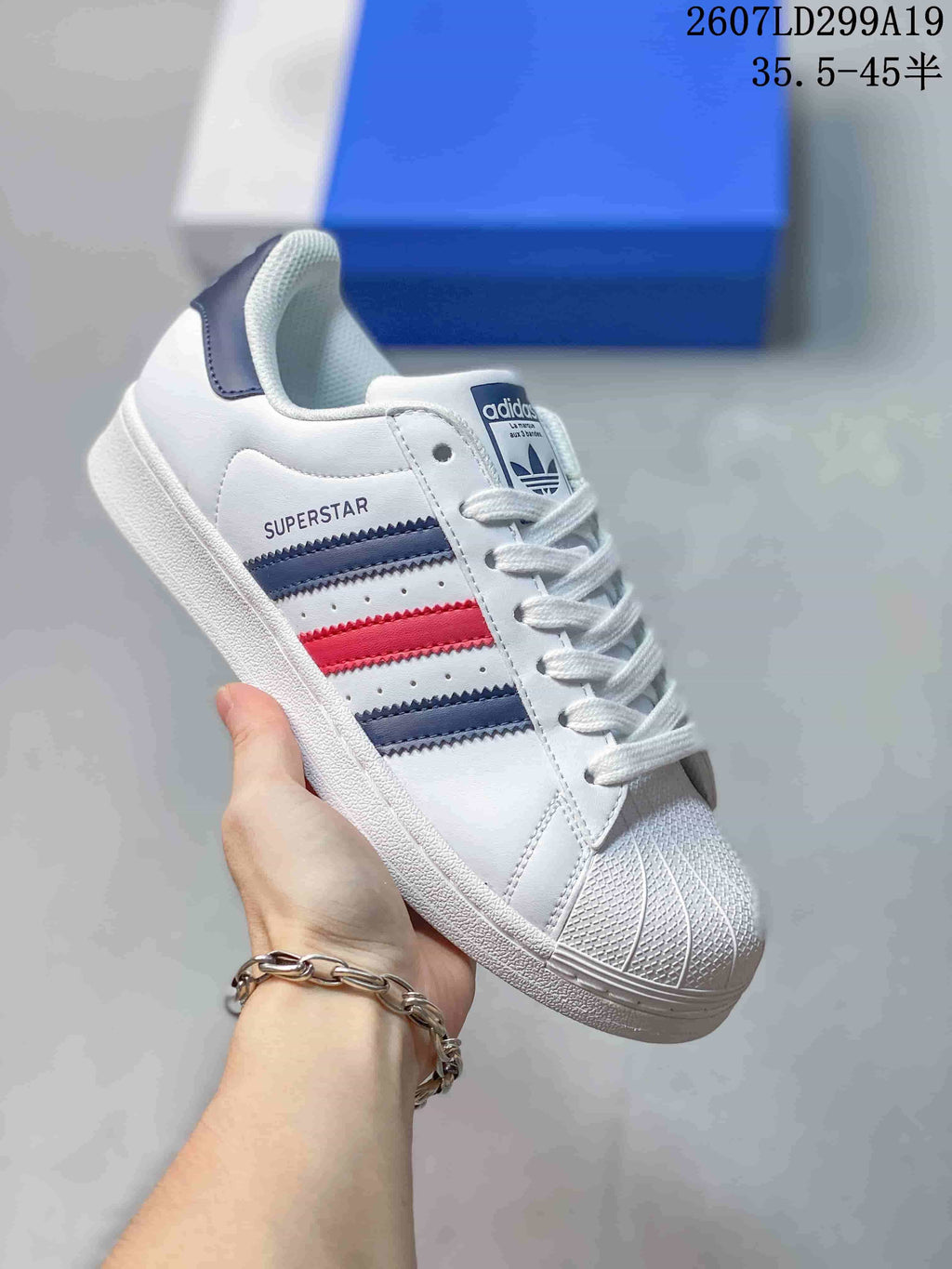 White adidas Superstar sneaker with navy and red serrated stripes and a blue trefoil tongue label.