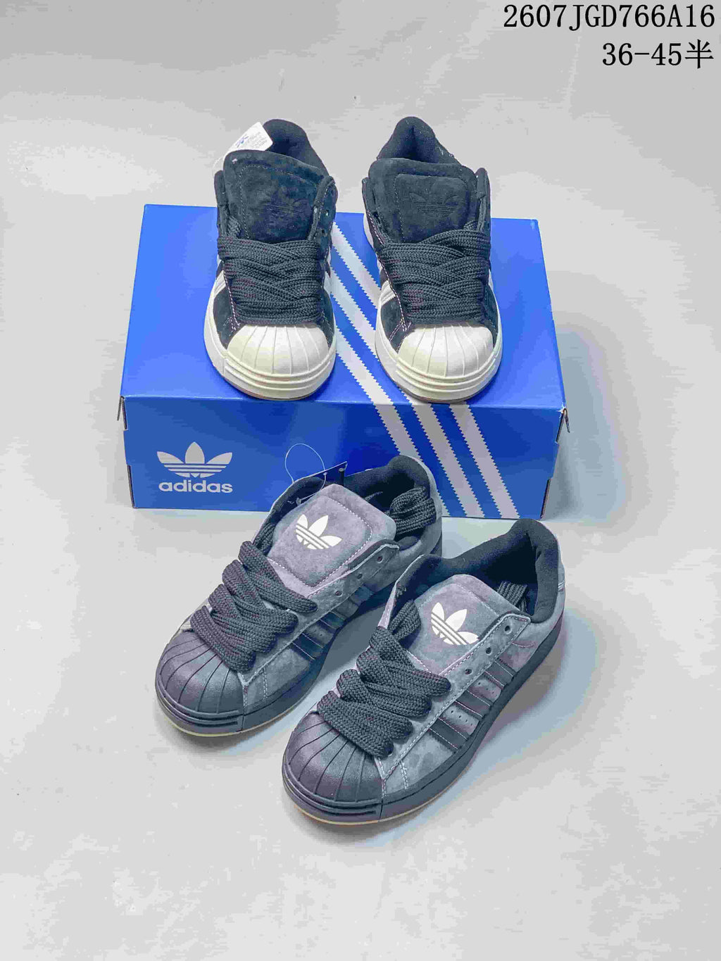 Front and overhead product view showing two pairs of dark shell-toe adidas sneakers with blue adidas packaging.