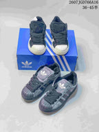 Front and overhead product view showing two pairs of dark shell-toe adidas sneakers with blue adidas packaging.