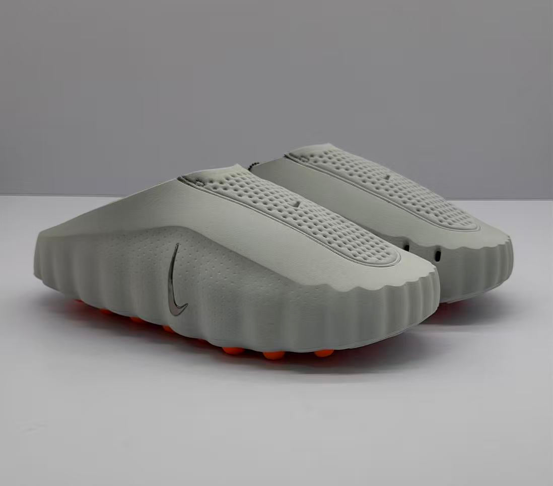 Three-quarter view of a pair of gray Nike slide sandals with perforated footbeds and orange outsole elements.