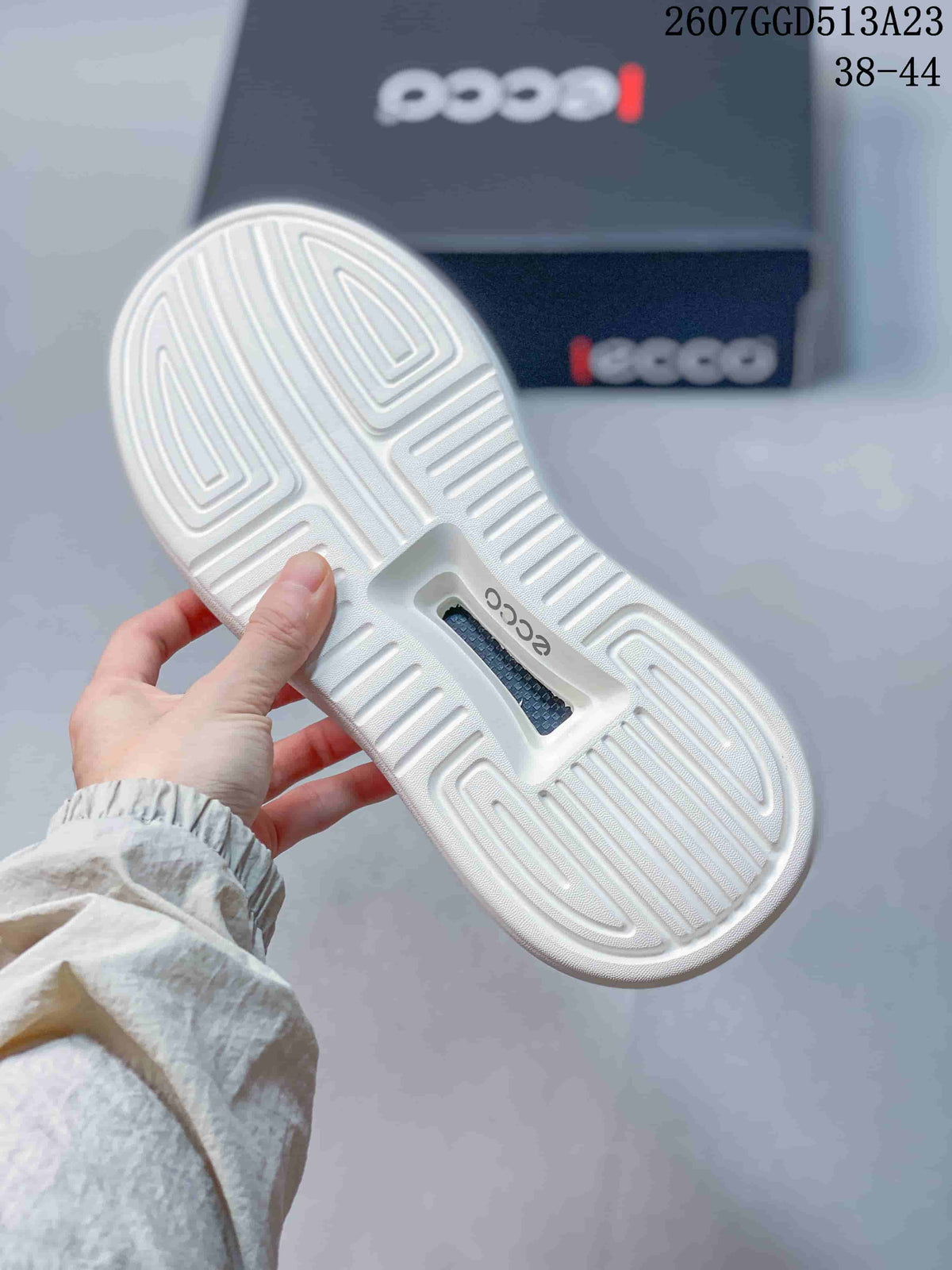 Underside of the white sneaker showing molded linear tread, a central ECCO logo detail, and the branded box in the background.