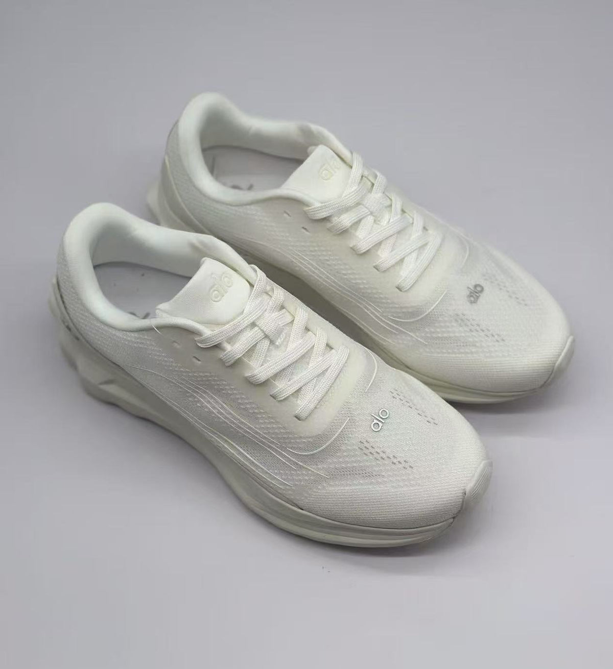 Top three-quarter view of a pair of white Alo sneakers with tonal laces and branding.