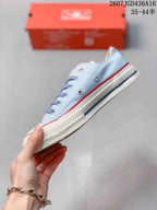 Side profile of the light blue low-top sneaker showing metal eyelets, white rubber midsole, and red and blue stripe detailing.