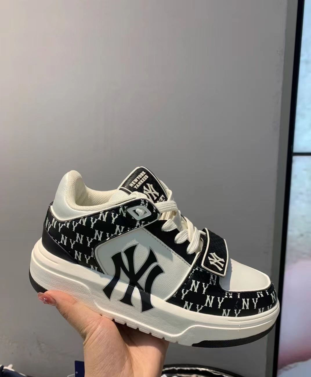 Black and white MLB M001 sneaker held in hand, showing New York Yankees graphics, a repeating monogram-style upper pattern, white laces, and a layered outsole.
