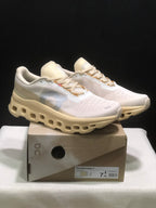 Pair of On Cloudmonster 1 sneakers in a light cream and white colorway displayed on the labeled shoe box.