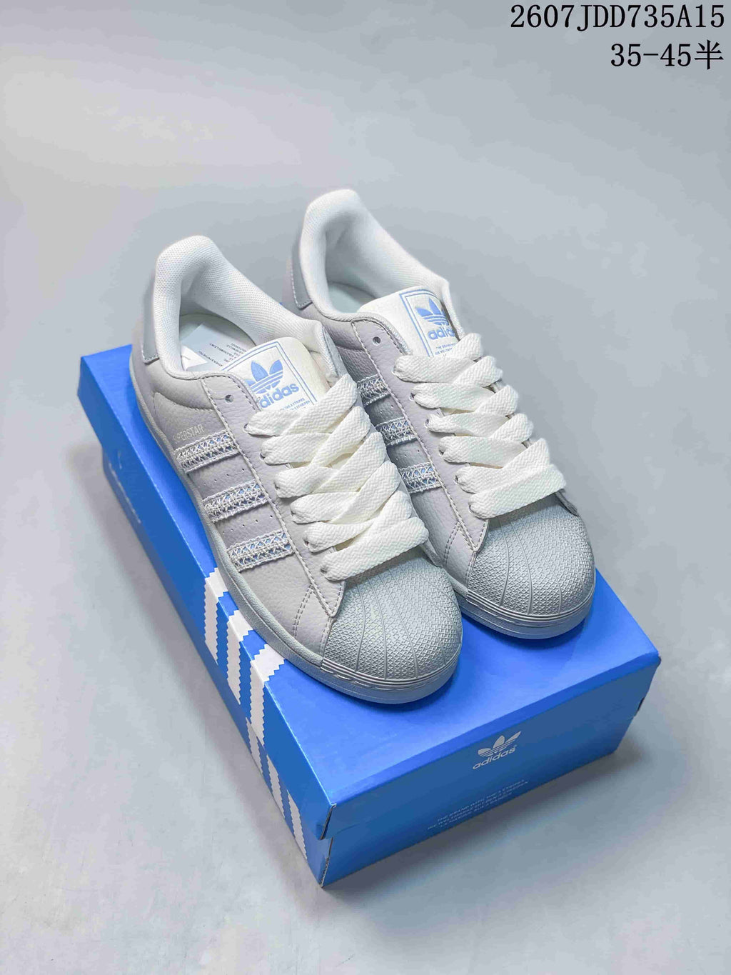 Pair of white and gray adidas Superstar sneakers displayed on a blue shoebox, showing the shell toes and blue branding.