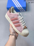 Angled side view of a pink adidas Superstar-style sneaker showing the shell toe, textured stripes, white laces and cream sole.
