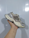 Side view of a beige and grey layered sneaker with mesh panels and a chunky segmented outsole, held against a light background.