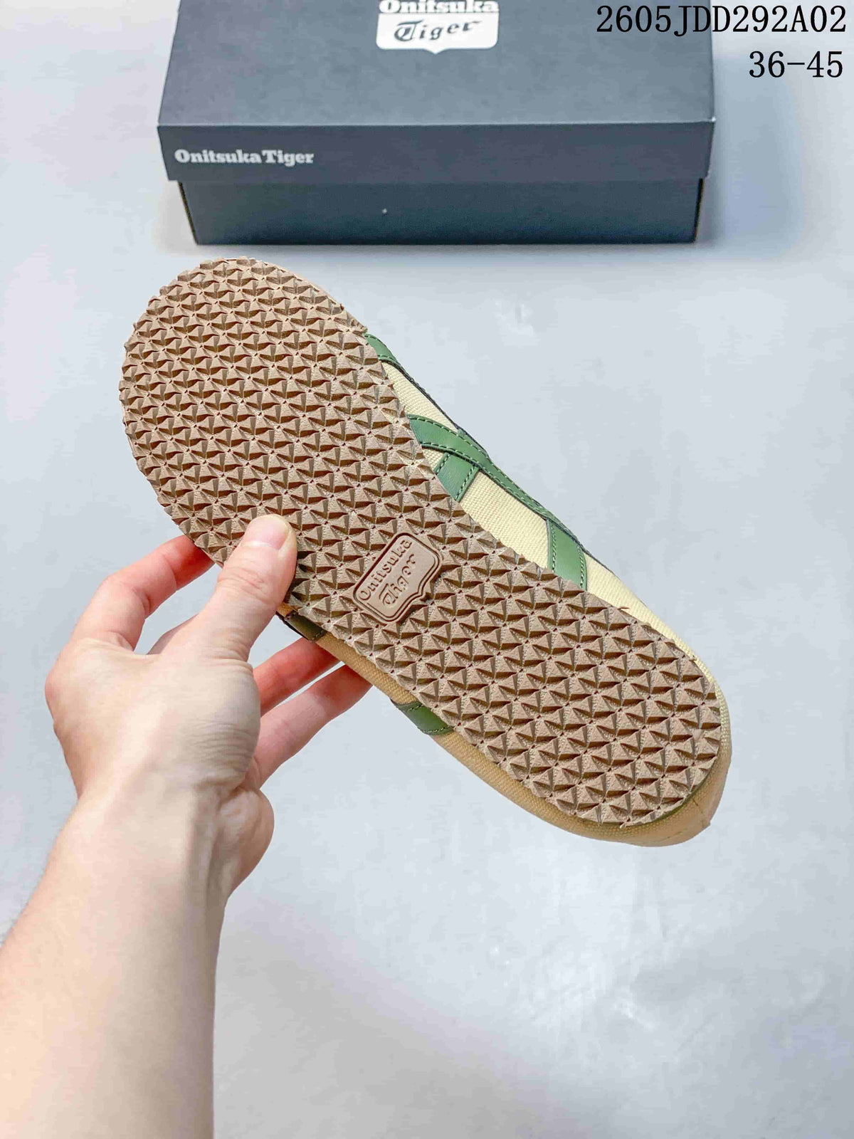 Underside view of an Onitsuka Tiger sneaker showing the repeating textured outsole pattern and branded outsole insert; a branded shoe box is in the background.
