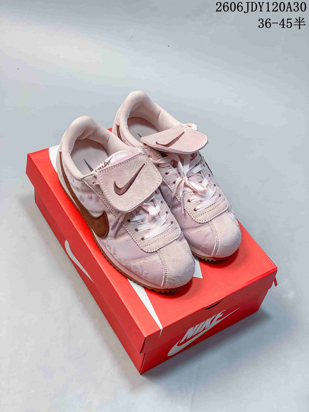 Pale pink low-top sneakers with brown Swooshes and flap details over the laces, photographed on a red shoe box.